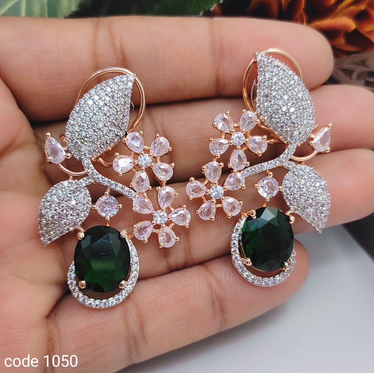 A pair of earrings featuring a central American diamond-like stone surrounded by smaller clear and green stones, set in a gold-colored mounting.