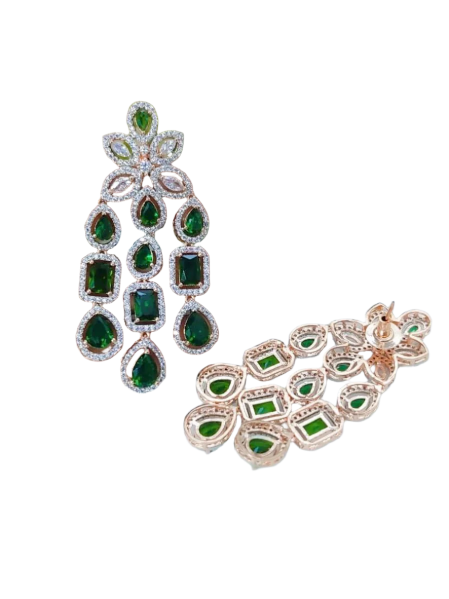 A pair of emerald-colored American diamond earrings with a metallic (potentially gold-plated) band, held in a hand against a floral background.