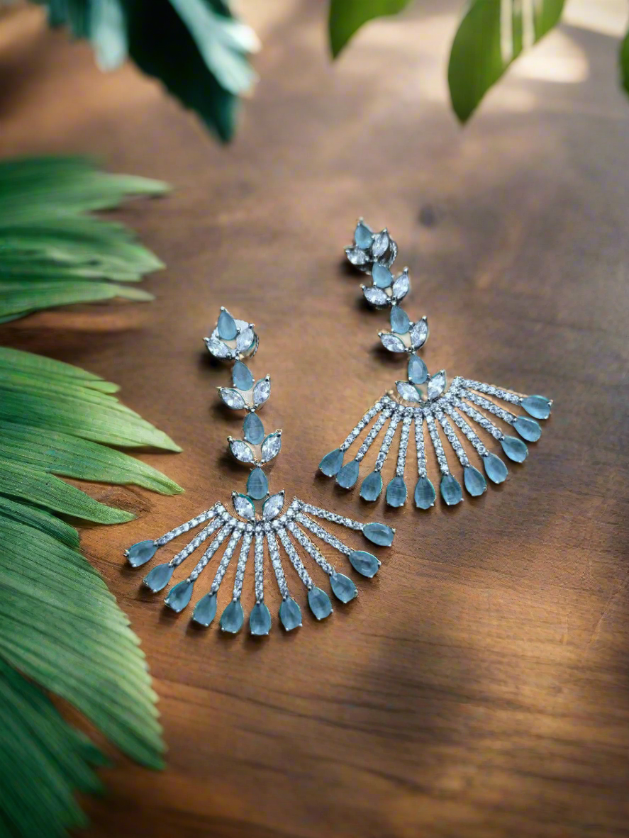 A pair of elegant blue and silver American diamond earrings with a chandelier design, displayed against a textured brown background.
